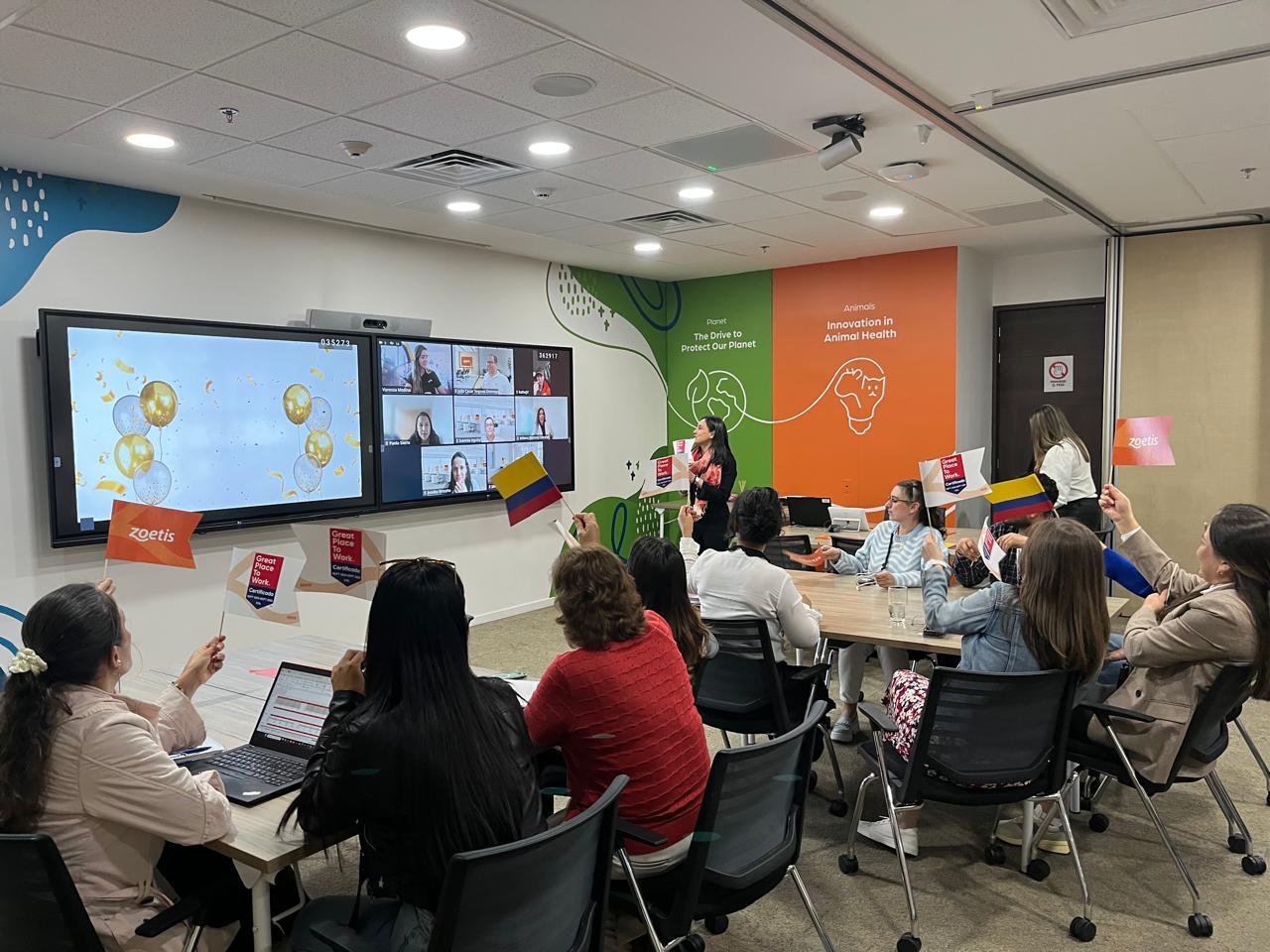 Alejandra dirigiendo una reunión de equipo, con colegas en la mesa y otros conectados en pantalla